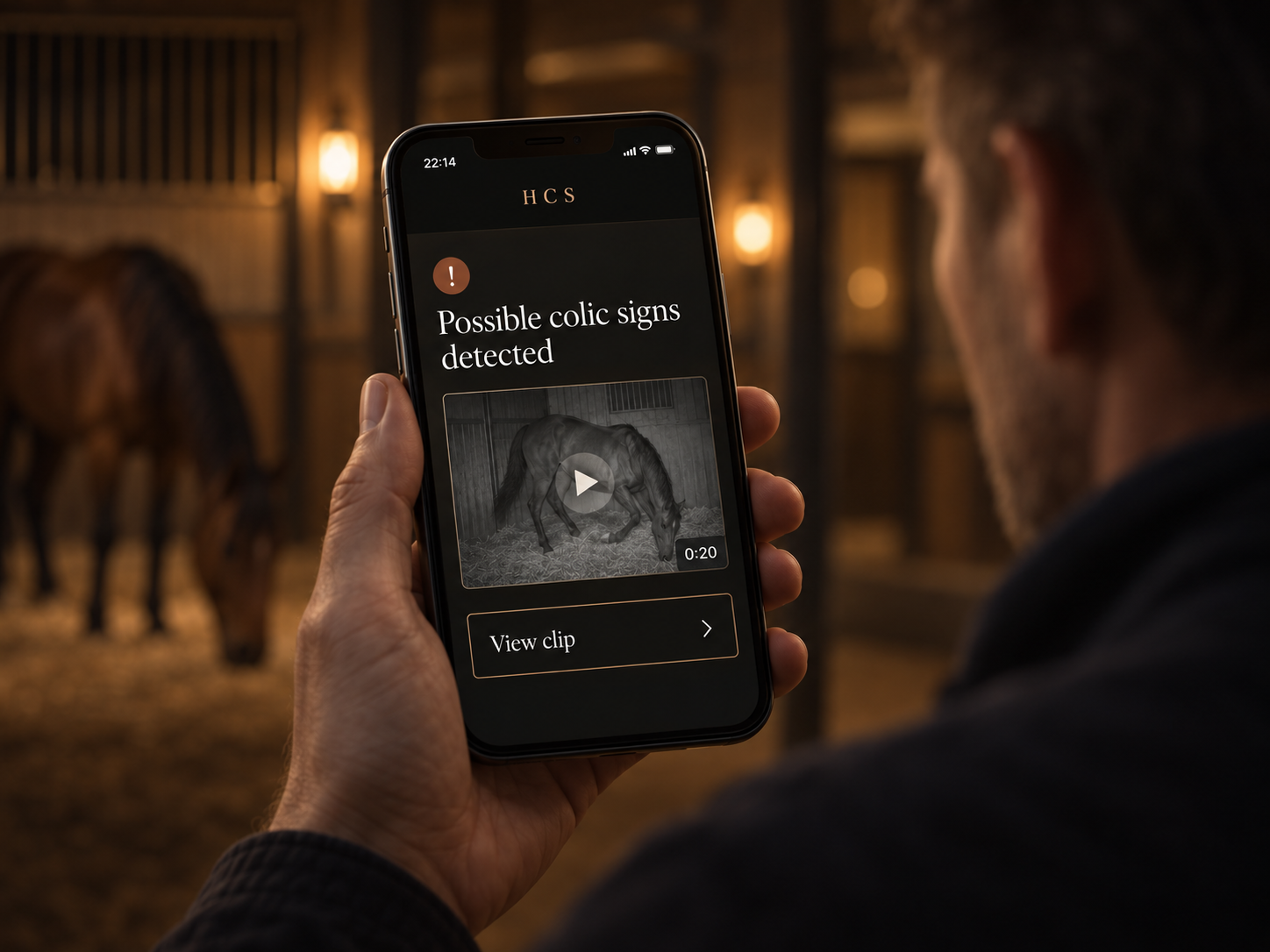 A man holds a phone showing an HCS alert with a horse clip in the stable.